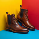 A pair of brown leather lace-up ankle boots with black elastic side panels, displayed on a bright blue surface against a split yellow and red background.