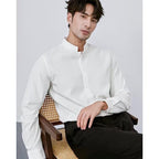 Man wearing a white shirt and black pants sitting on a wooden chair against a plain background