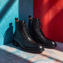A pair of black leather lace-up ankle boots with side zippers, displayed on a textured surface between a blue wall on the left and a red wall on the right, with dramatic shadows cast across the scene.