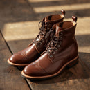 A pair of brown leather lace-up ankle boots with brogue detailing on the toes, displayed on a rustic wooden surface with warm sunlight and shadows.