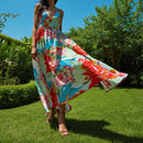 A woman wearing a flowing maxi dress with a colorful floral print, standing on a green lawn with trees and blue sky in the background.